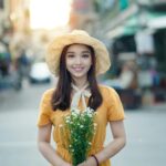 Smiling woman in a yellow dress and straw hat holding flowers on a sunlit street.